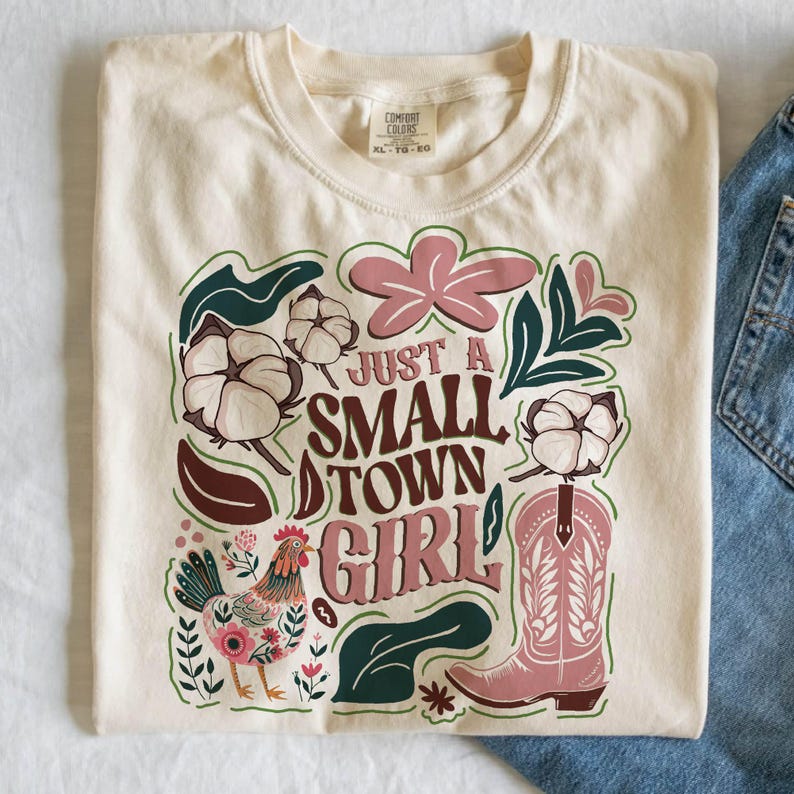 Pu&ograve; includere: T-shirt color crema con la scritta "Just a Small Town Girl" in un carattere decorativo. Il design include uno stivale da cowboy rosa, fiori di cotone, un gallo e accenti floreali. La maglietta &egrave; piegata.