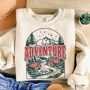 May include: A cream-colored sweatshirt with a vintage-style graphic of a campfire scene. The graphic features a mountain range, two red tents, a campfire, and trees. The text "Seek Adventure" is written in red and black letters.