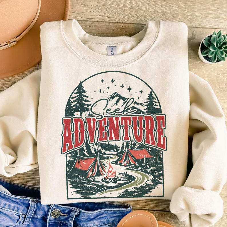 May include: A cream-colored sweatshirt with a vintage-style graphic of a campfire scene. The graphic features a mountain range, two red tents, a campfire, and trees. The text "Seek Adventure" is written in red and black letters.