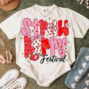 May include: White t-shirt with a pink and red graphic that says "Strawberry Festival". The graphic is made up of different patterned squares and includes strawberries and flowers.