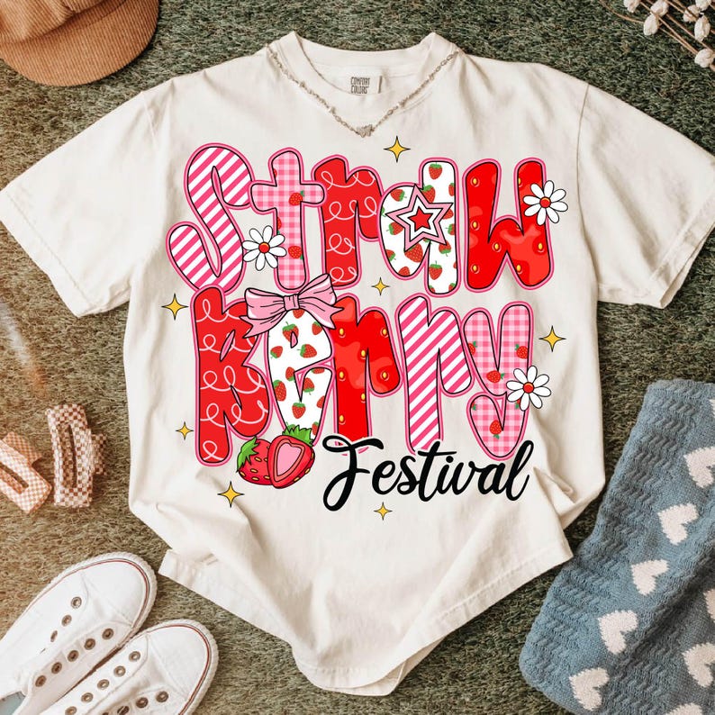 May include: White t-shirt with a pink and red graphic that says "Strawberry Festival". The graphic is made up of different patterned squares and includes strawberries and flowers.