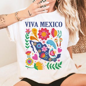 May include: White t-shirt with a colorful floral design and the text "Viva Mexico" printed on the front. The design features a silhouette of Mexico with flowers, leaves, and hearts.