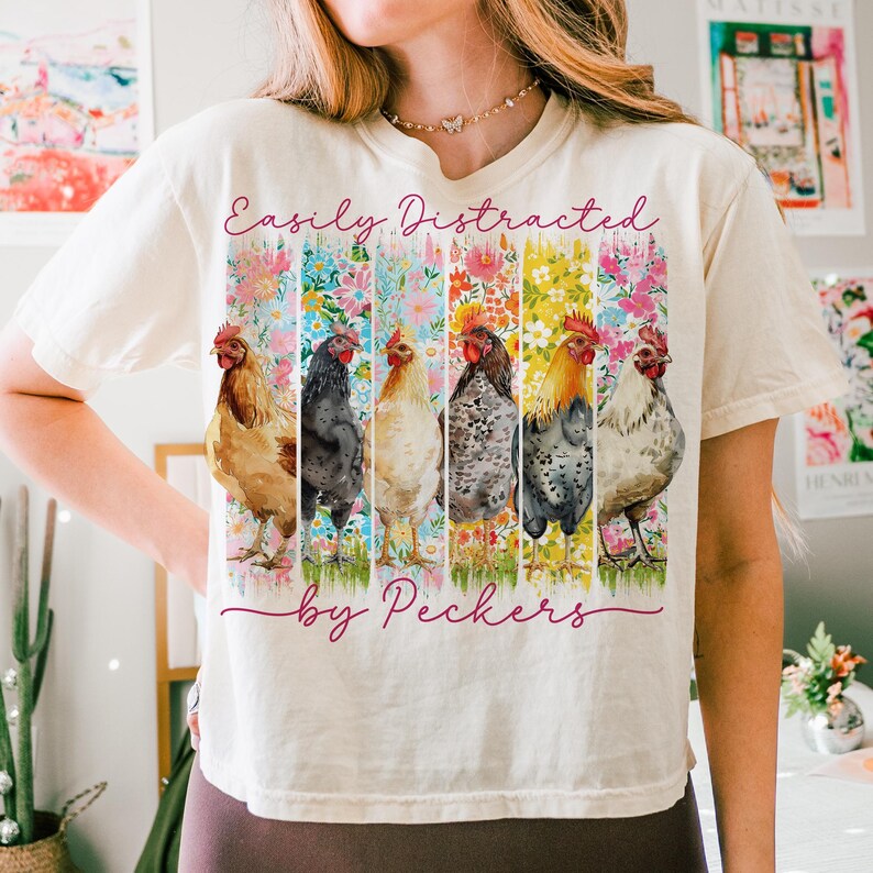 May include: Cream-colored t-shirt featuring a colorful graphic of six chickens and the text "Easily Distracted by Peckers." The design includes floral patterns and a whimsical font, perfect for a casual, fun look.