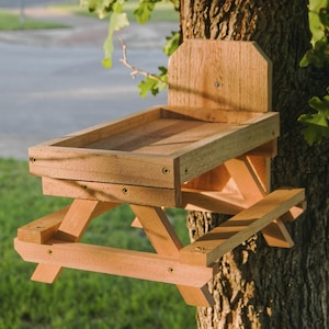 May include: A wooden picnic table-style bird feeder attached to a tree. The feeder has a rectangular tray and a backboard. The design includes a miniature picnic table with attached benches, perfect for attracting birds to your yard.