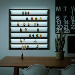 Op de afbeelding: Een grijs wandrek met een wit licht erachter, waarop een verzameling kleine, kleurrijke Lego-minifiguren wordt getoond. Het rek heeft zeven planken, elk met een andere minifiguur. De minifiguren zijn allemaal in verschillende kleuren en hebben verschillende accessoires. Er is een kalender aan de muur achter het rek, en een plant in een bruine pot op een tafel voor het rek.