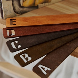 May include: Five wooden bookmarks with letters A through E cut out. The bookmarks are stained in different shades of brown, from light to dark.