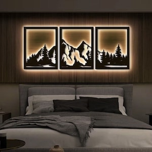 Può includere: Un'opera d'arte da parete in metallo nero che rappresenta una catena montuosa con alberi di fronte. L'opera è illuminata da dietro, creando un bagliore caldo. L'opera è montata su una parete in legno scuro sopra un letto con biancheria da letto grigia.