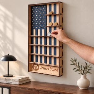 Op de afbeelding: Een rechthoekige houten vitrine met een gestileerd Amerikaans vlagontwerp met blauwe strepen en sterren. De kast heeft horizontale planken voor het bewaren van challenge coins. Het onderste gedeelte is gegraveerd met James Stone en een Navy-zegel.