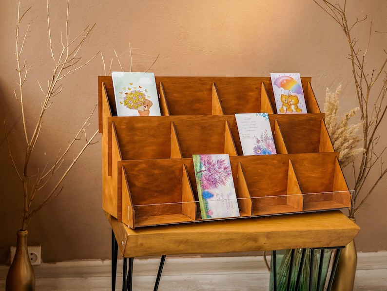 May include: A wooden card display stand with 12 compartments, each holding a greeting card with a different design. The stand is made of natural wood and has a clear acrylic front.