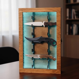 Könnte beinhalten: Eine Holzvitrine mit drei Messern. Die Messer haben unterschiedliche Klingenformen und Griffdesigns. Die Vitrine hat einen braunen Holzrahmen und einen blau-grün beleuchteten Hintergrund. Die Messer werden von schwarzen Halterungen gehalten.