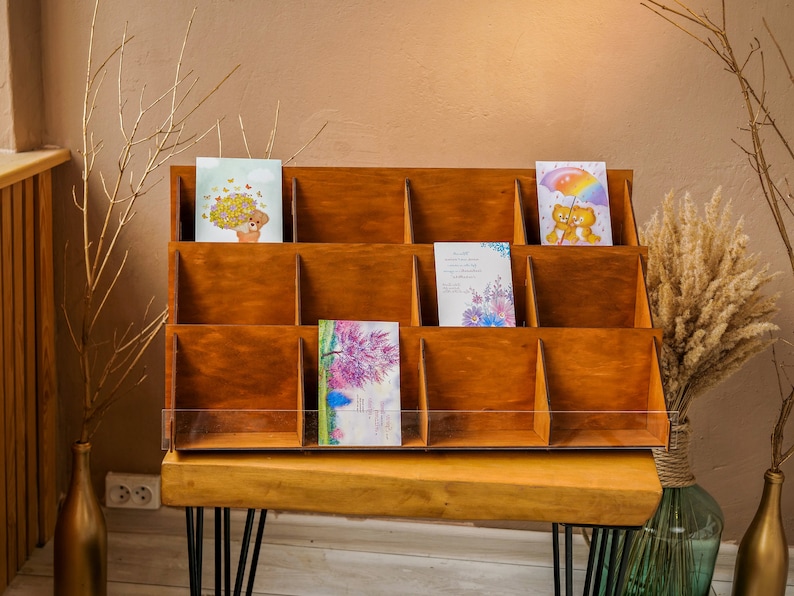 May include: A wooden card display stand with 12 compartments, each holding a greeting card with various designs. The stand is on a wooden table with black metal legs.