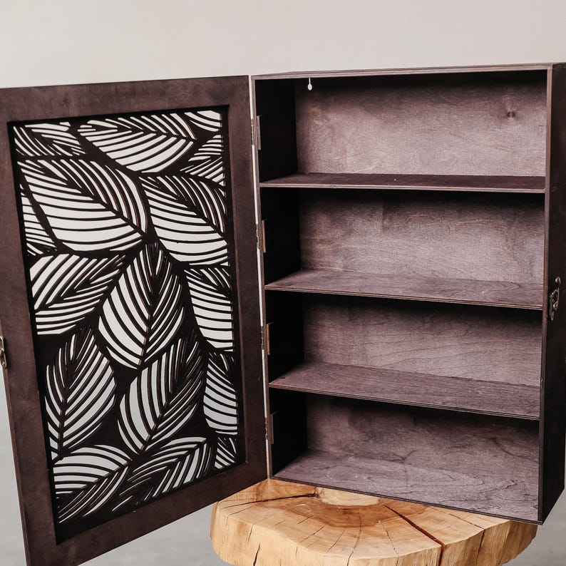 May include: A dark brown wooden cabinet with a door featuring a white leaf pattern. The cabinet has three shelves inside.