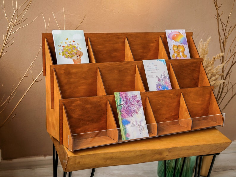 May include: A wooden card display stand with 12 compartments for holding greeting cards. The stand is made of natural wood and has a clear acrylic front panel. The stand is sitting on a wooden table.