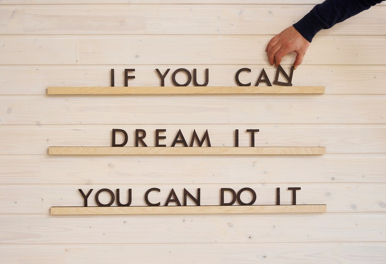 May include: A motivational quote, "If you can dream it, you can do it", is displayed on three wooden shelves against a white wood background. The quote is spelled out using individual black letters.