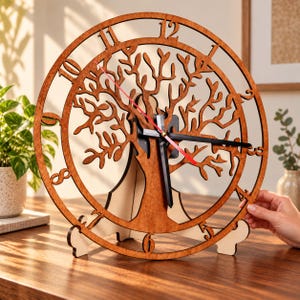 May include: A circular wooden wall clock featuring a laser-cut tree silhouette in the center. The clock face has cutout numbers from one to twelve along the outer ring. It includes black hour and minute hands and a thin red second hand.
