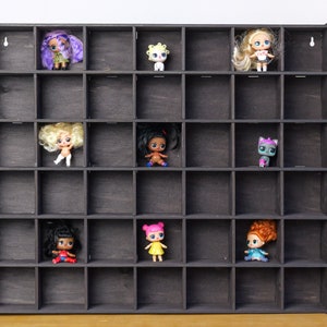 May include: A dark brown wooden display shelf featuring a grid of thirty square compartments. Eight small collectible dolls with various hair colors and outfits are positioned within individual cubbies, leaving the remaining spaces empty.