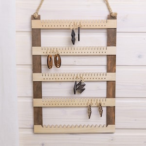 Könnte beinhalten: Holz-Ohrringhalter mit vier Reihen von Stiften. Der Halter ist braun gebeizt und hat eine helle Holzoptik. An den Stiften hängen Ohrringe, darunter geometrische Formen und Holzringe.