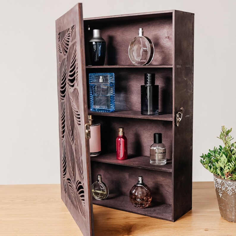 May include: A dark brown wooden cabinet with a carved leaf design on the door. The cabinet has four shelves and is filled with various perfume bottles. The door is open and the shelves are visible.
