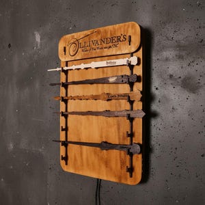 Könnte beinhalten: Holz-Zauberstab-Display mit sechs Zauberstäben. Das Display besteht aus hellbraunem Holz und trägt den Text "Ollivander's Makers of Fine Wands since 382 BC". Jeder Zauberstab hat ein einzigartiges Design und Farbe.