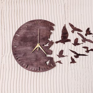 Puede incluir: Un reloj de madera marrón con una aguja de hora y minutos dorada. La esfera del reloj está parcialmente ausente, con una bandada de pájaros volando fuera de la abertura. Los pájaros están recortados en la madera y están en silueta.