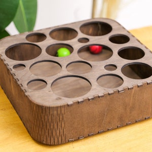 May include: A wooden box with a square base and rounded edges. The box has multiple circular cutouts of varying sizes. There are two small balls inside the box, one green and one red.