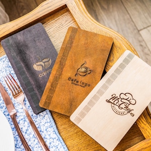 May include: Three wooden menu covers with different designs. The first cover is black with a coffee cup logo and the text "Coffee". The second cover is brown with a coffee cup logo and the text "Cafe logo, slogan here". The third cover is light brown with a chef hat logo and the text "Mr. Chef Restaurant".