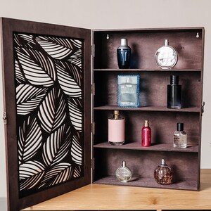 May include: A wooden cabinet with a leaf-patterned door, open to reveal shelves filled with various perfume bottles. The bottles are in different shapes and sizes, and some have labels.