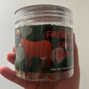 May include: A clear plastic jar with a screw-on lid, containing freeze-dried beef. The label features a cartoon cow and the words "Freeze Dried Grass Fed Beef Livers". The text "Raw Diet Friendly Dogs" is also visible.