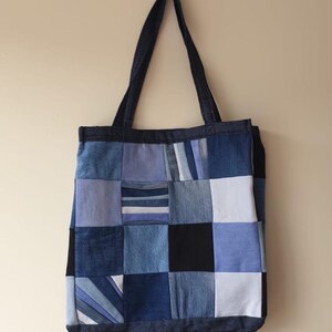 May include: A tote bag constructed from a patchwork of various blue and black fabric squares. The design features denim and cotton textures in shades ranging from light sky blue to deep navy and black, with two long shoulder straps at the top.