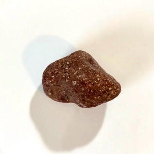 Pode incluir: Uma rocha castanho-avermelhada com uma superfície áspera e texturizada e pequenas manchas brancas. A rocha tem uma forma irregular e parece ser uma pedra natural. A imagem é tirada contra um fundo branco.