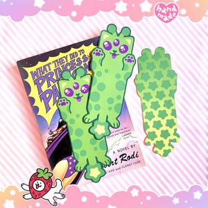 May include: Green alien cat bookmarks with purple eyes and spots, next to a book titled "WHAT THEY DID TO PRINCESS PA". One bookmark is a star-shaped design. The image has a pink and white striped background.