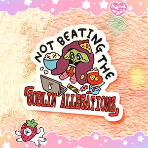 May include: A white sticker with a cartoon goblin character surrounded by snacks and a laptop. The text reads "NOT BEATING THE GOBLIN ALLEGATIONS" in black and red. A small "hand made" logo is in the top right corner.