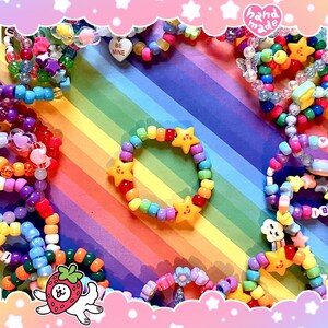 May include: A selection of colourful beaded bracelets and necklaces. One bracelet has yellow star charms. The background is a rainbow gradient. Some pieces have heart-shaped beads and the words "BE MINE". The image has a "hand made" stamp.