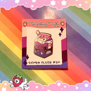 Op de afbeelding: Een kleurrijke emaille pin in de vorm van een melkpak, met de tekst "Genda Fluid Pin". De pin heeft een roze, paars en blauw kleurenschema met een rietje en steraccenten. De pin zit op een kaart met de tekst "Delightfully Dinky".