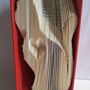 May include: A book art sculpture of a heart shape, folded from the pages of a book, displayed in a red book stand with a red ribbon.