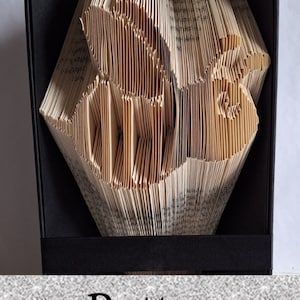 May include: A book art sculpture of a bird, folded from the pages of a book. The sculpture is displayed in a black frame with a black base. The word "Pattern" is printed on a gray textured surface below the frame.