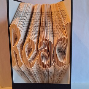 Könnte beinhalten: Eine Buchkunstskulptur des Wortes "Read", die durch das Falten der Seiten eines Buches entstanden ist. Die Skulptur steht auf einem schwarzen Sockel.