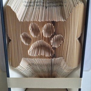 May include: A book folded into the shape of a paw print. The book is bound in black and has a cream-colored ribbon bookmark. The book is displayed on a white surface with the word "Pattern" printed below it.