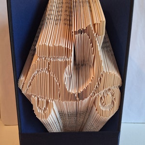 May include: A book art sculpture of a car made from the pages of a book. The book is bound in a dark blue cover and is displayed on a stand.