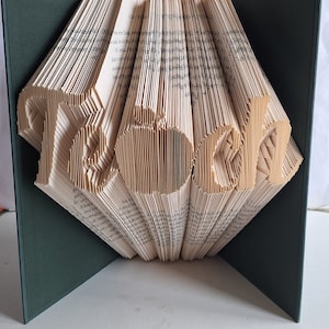 May include: A folded book sculpture with the word "Read" and an apple shape cut into the pages. The book is open, revealing the text of the pages, and is set against a dark green background.