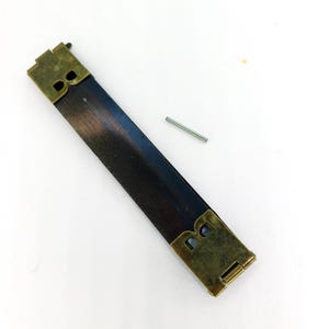 May include: A brass-colored metal strap with a dark brown leather-like center. The strap has two metal clasps on each end and a small metal pin.