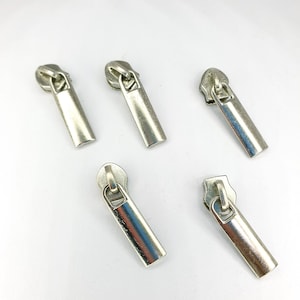 May include: Five silver zipper pulls with different styles. The pulls are all metal and have a shiny finish.