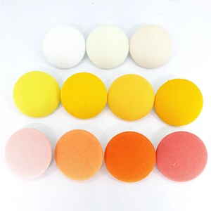 May include: A set of 10 round fabric buttons in a gradient of colours, starting with white and transitioning to pink. The buttons are arranged in three rows, with three buttons in the top row, four buttons in the middle row, and three buttons in the bottom row.