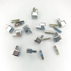 Peut inclure: Un ensemble de coins de fixation en métal argenté, certains avec une petite lettre 'M' imprimée dessus. Les coins sont dispersés sur une surface blanche.