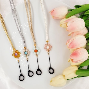 Könnte beinhalten: Vier geflochtene Brillenketten mit Blumenanhängern. Die Ketten sind in Silber, Beige, Weiß und Gold. Die Blumen sind aus bunten Perlen gefertigt.