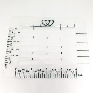 Könnte beinhalten: Ein transparentes Lineal aus Kunststoff mit einem Herzdesign oben. Das Lineal ist in Zoll und 1/4 Zoll unterteilt. Das Lineal ist 10 cm lang.
