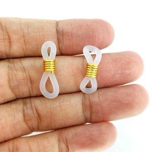 Peut inclure: Deux supports de cordon pour lunettes en plastique blanc avec des accents en métal doré. Les supports sont conçus pour être fixés aux extrémités des montures de lunettes.