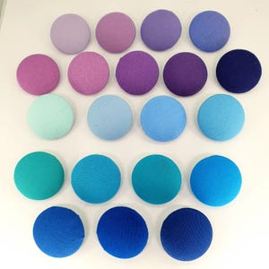Puede incluir: Una colección de 20 botones redondos de tela en tonos de morado, azul y turquesa. Los botones están dispuestos en un patrón de degradado, comenzando con el tono más claro de morado en la parte superior y terminando con el tono más oscuro de turquesa en la parte inferior.