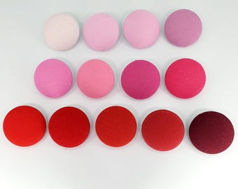 May include: A set of 12 round fabric buttons in shades of pink and red. The buttons are arranged in three rows of four, with the lightest shade at the top and the darkest shade at the bottom.