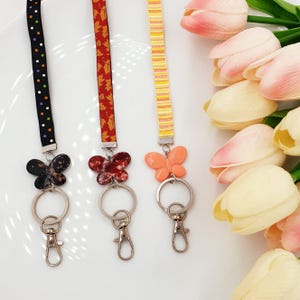 May include: Three colourful lanyards with butterfly charms and silver keyrings. The lanyards are black with white polka dots, red with autumn leaves, and yellow with orange stripes.